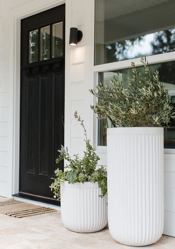 a modern porch with a black door, a duo of large fluted planters with greenery and a woven rug plus a black spotlight