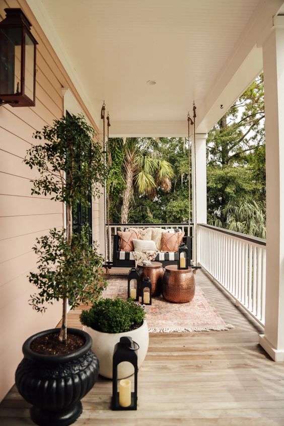 a modern porch with a hanging black bench with upholstery and pillows, metal side tables, candle lanterns and potted greenery