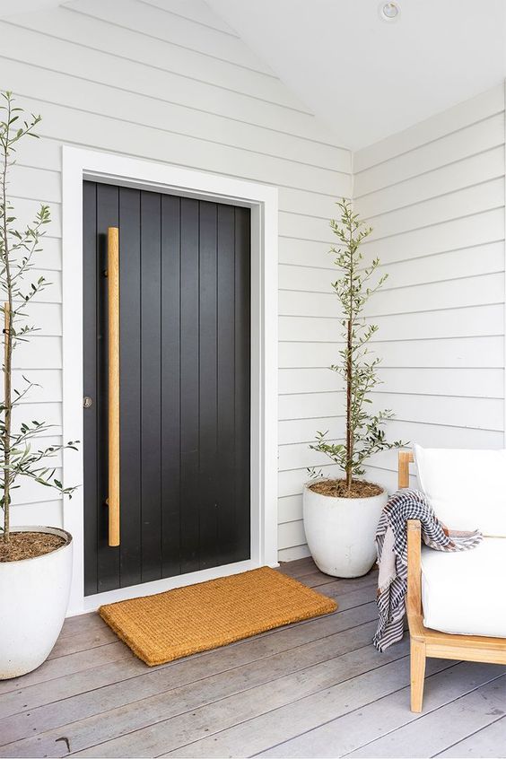 a modern porch with white shiplap on the walls and a black door, a white chair, potted plants and a woven rug