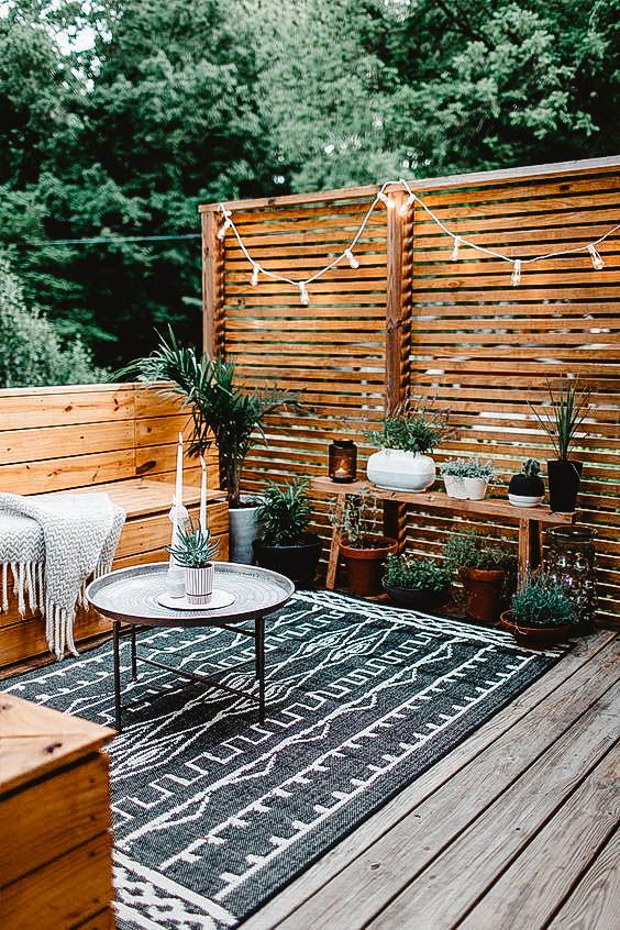 a modern rustic terrace with a wooden deck, built-in benches with a blanket, a bench with potted plants and some lights over the space