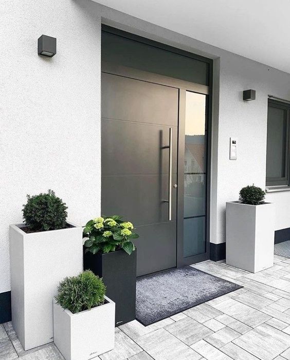 a modern to minimalist front porch with neutral and black square planters with greenery, a heavy black door and a rug
