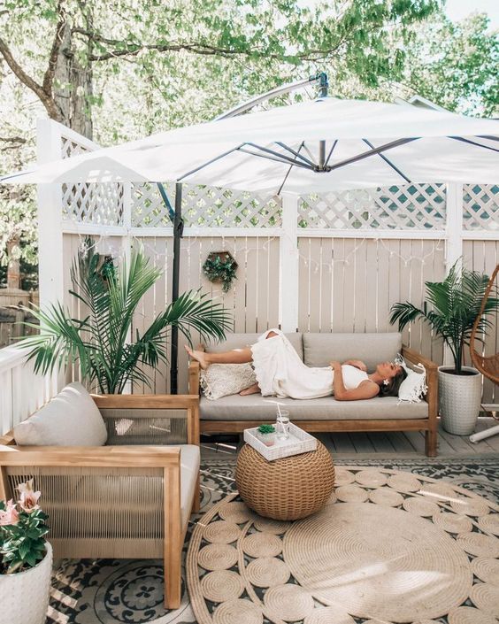 a modern tropical patio with wooden furniture, a woven side table, potted plants, layered rugs and an umbrella over the space