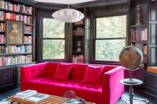 a moody and elegant black home library with lots of bookshelves, a hot pink sofa and a printed rug, a globe and a pendant lamp