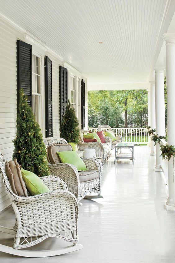 a neutral farmhouse porch with white wicker furniture, neutral and green pillows, some greenery and Christmas trees