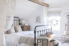 a neutral modern country bedroom with white planked walls, a wooden beam over the metal bed, neutral furniture and a large candle lantern