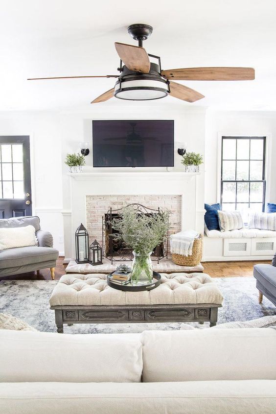 a neutral modern farmhouse living room with a fireplace clad with brick, neutral furniture and built-in daybeds, an upholstered ottoman and a fan