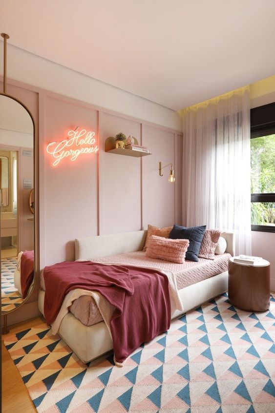 a pale pink paneled accent wall, a creamy daybed with comfy pillows, a neon sign, a large mirror and a round side table