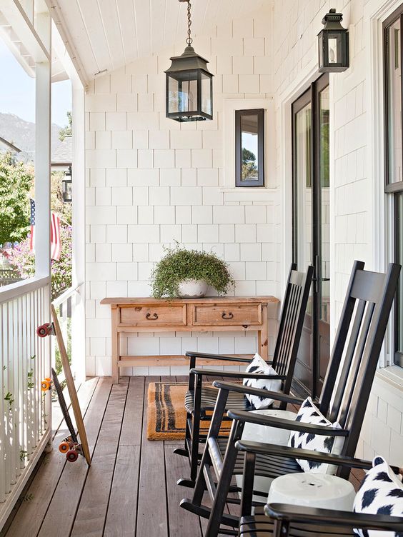 a pretty and simple farmhouse porch with a vintage console table, black rockers and printed pillows, potted greenery and some lanterns is welcoming