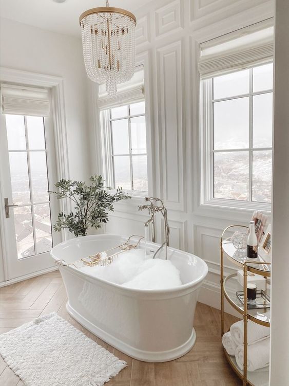 a refined and chic fancy bathroom with white paneled walls, windows, a luxurious bathtub, a crystal chandelier and a potted plant