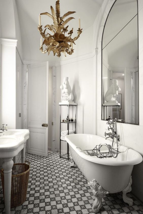 a refined and luxurious bathroom with white walls, a mosaic tile floor, a white vintage tub and an oversized mirror and a gold chandelier