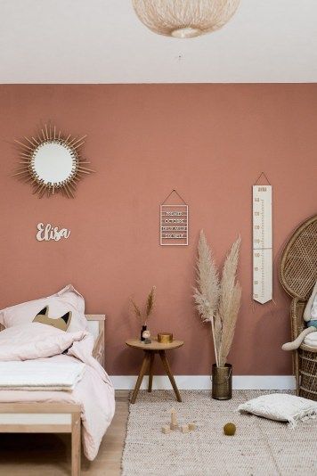 a small boho kid's bedroom with a pink accent wall, simple wooden furniture and a peacock chair, a sunburst mirror and pampas grass