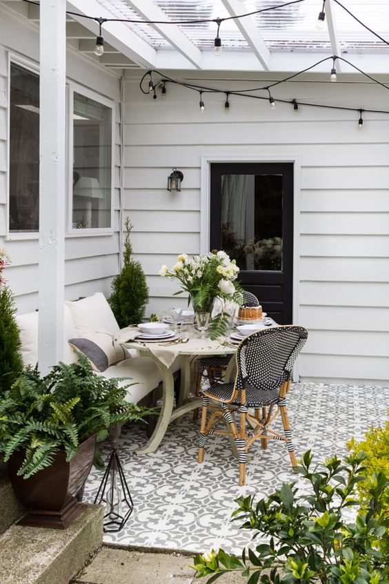 a small modern farmhouse terrace with mosaic tiles, vintage and modern wooden and rattan furniture, potted plants and blooms and lights over the space