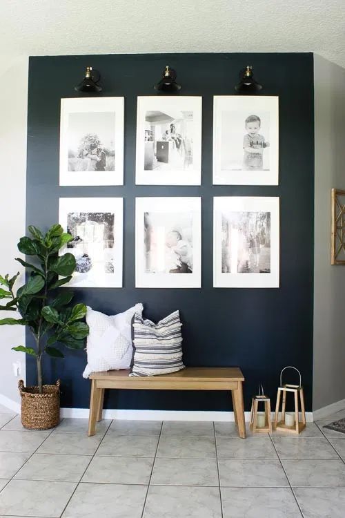 a stylish entryway with a navy accent wall, a bench with pillows, wooden candle lanterns, a black and white grid gallery wall and a potted plant