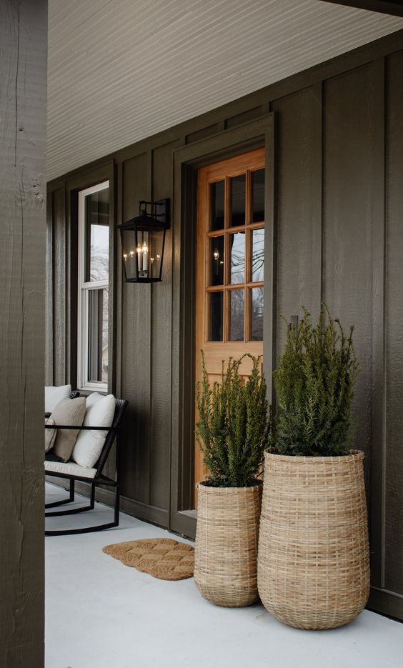 a stylish modern front porch with black trim walls, woven planters with greenery, a woven rug, a white upholstered chair