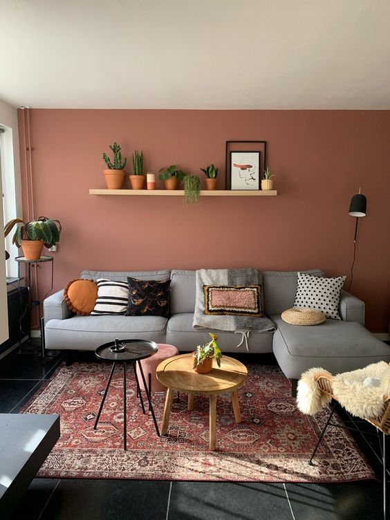 a stylish modern living room with a pink accent wall, a grey sectional, a printed boho rug, a shelf with lots of potted plants and an arrangement of side tables