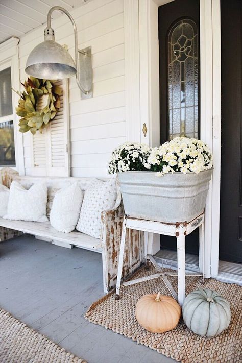 a vintage farmhouse porch with a white bench and printed pillows, a vintage metal sconce, a leaf wreath, potted blooms and pumpkins