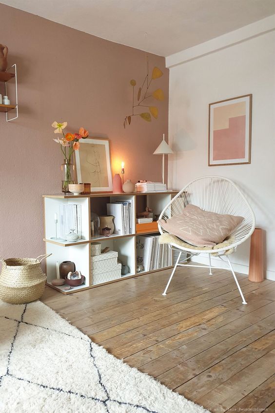 a welcoming Scandinavian nook with an open storage unit, a white chair, some lamps, a pale pink accent wall and a basket