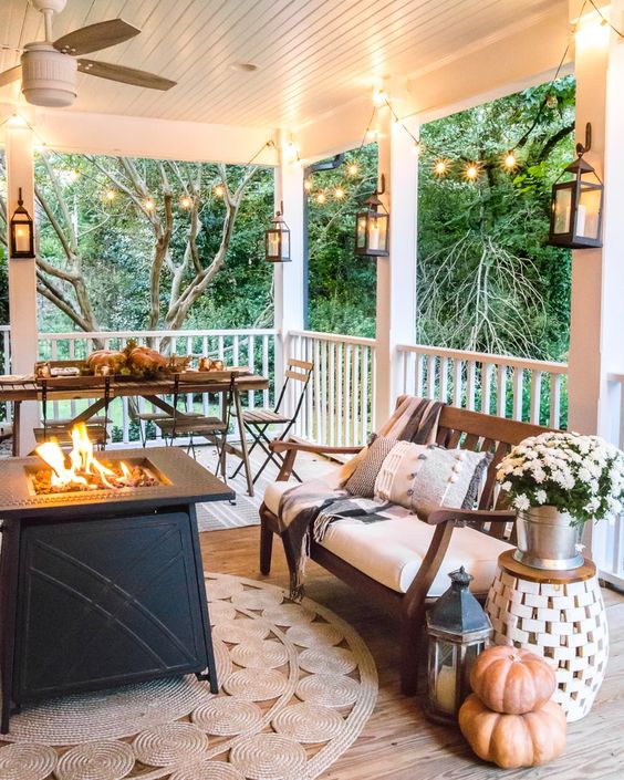 a welcoming farmhouse porch with a fire pit, a dining set, a wooden bench with upholstery, pumpkins and potted blooms in a bucket