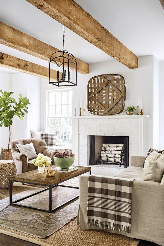a welcoming modern country living room with a brick clad fireplace, neutral furniture, layered rugs, printed textiles and wooden beams on the ceiling