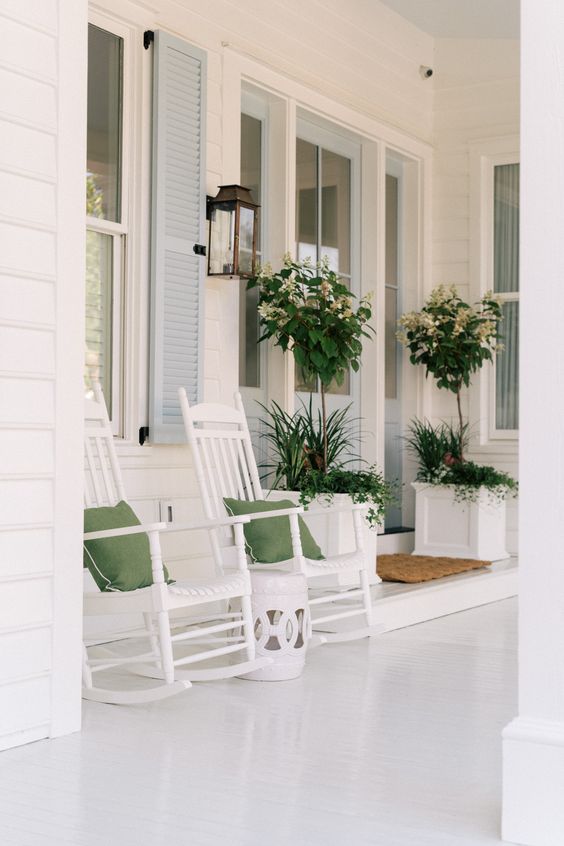 a white farmhouse porch with rockers and green pillows, a side table, a lantern and lots of greenery in large white planters is chic and clean