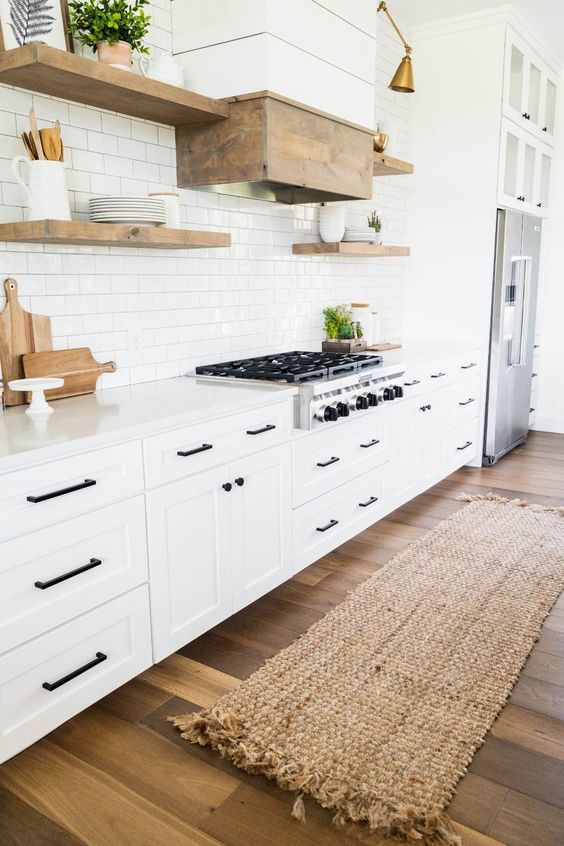 a white modern country kitchen with shaker style cabinets, white stone countertops, wooden floating shelves and a wooden hood