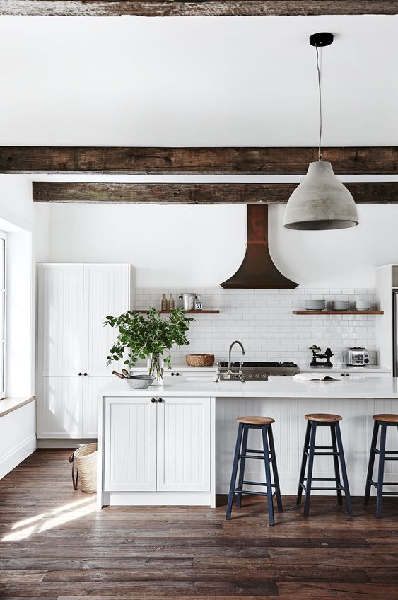 a white modern country kitchen with white planked cabinets, dark ceiling beams, pendant lamps, tall stools and shelves