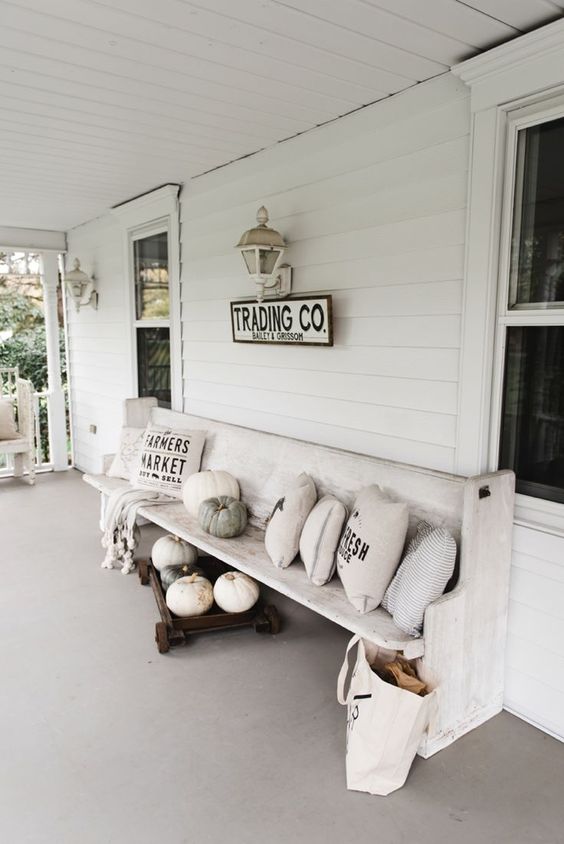a white vintage farmhouse porch with neutral pillows, pumpkins, lanterns and a bag with firewood is styled for the fall in a lovely way