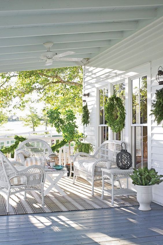 an elegant farmhouse porch with white wicker furniture, striped textiles, potted greenery and evergreens is very welcoming