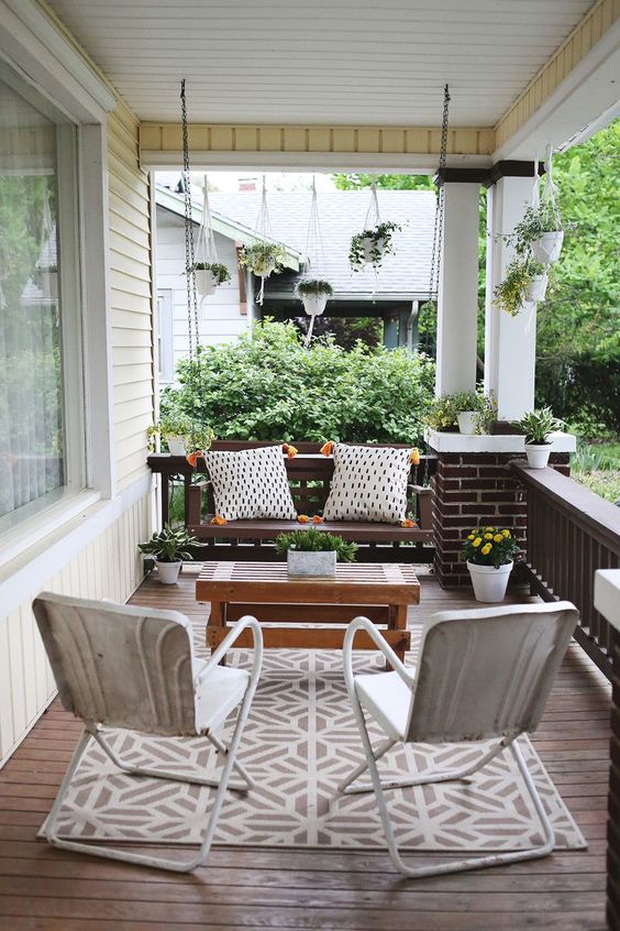 an elegant modern farmhouse porch with a dark stained suspended bench and pillows, white chairs, a wooden coffee table and potted greenery and blooms