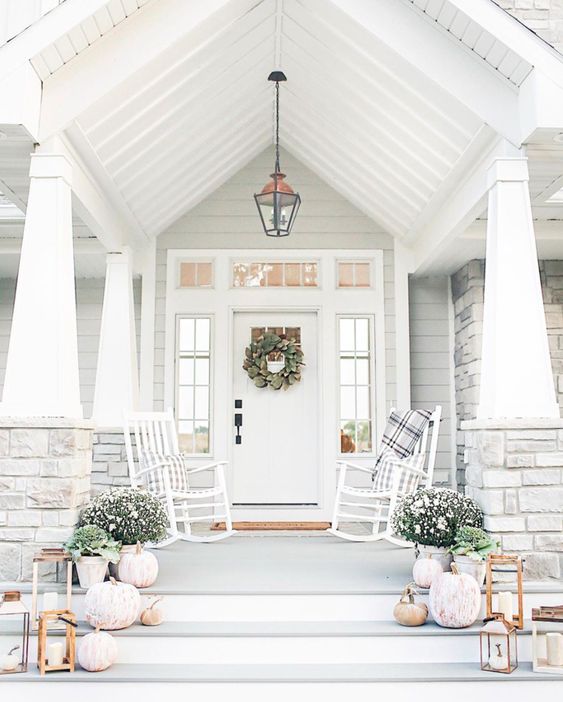 an elegant white farmhouse porch with a couple of white rockers and plaid textiles, potted blooms, candle lanterns and whitewashed pumpkins is amazing