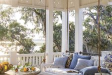 an inviting farmhouse porch with a hanging upholstered bench, a large round table, a rattan pouf and lovely textiles in blues
