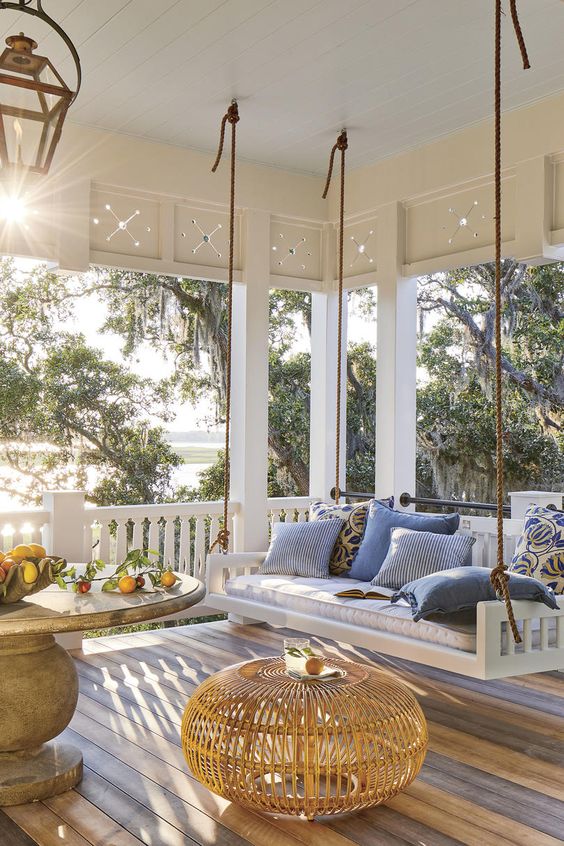 an inviting farmhouse porch with a hanging upholstered bench, a large round table, a rattan pouf and lovely textiles in blues