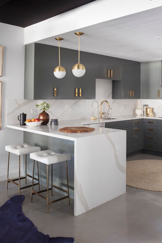 a refined kitchen with grey cabinets, a large kitchen island with a waterfall countertop, brass fixtures and gold pendant lamps