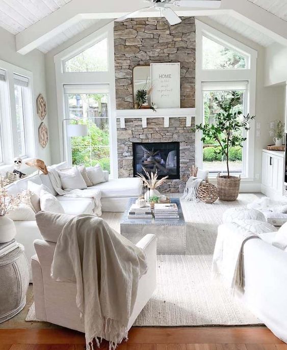 a white farmhouse living room with a fireplace clad with stone, white furniture, potted plants, neutral textules and views