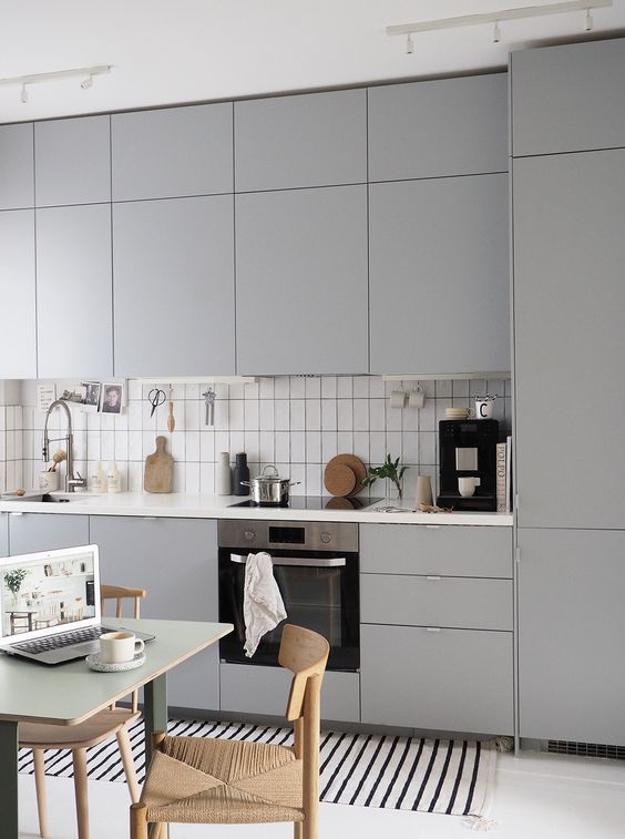 a modern grey kitchen with sleek cabinets, a white tile backsplash, a small table and woven chairs plus a striped rug