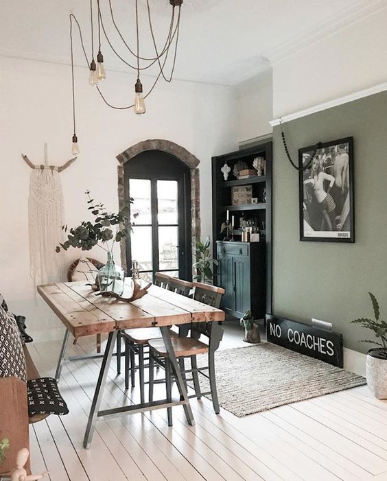 a boho dining room with a sage green accent wall, a black buffet, a trestle dining table, wooden chairs and bulbs plus macrame