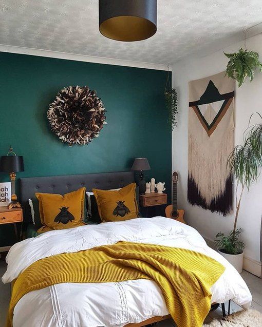 a bold boho bedroom with a dark green accent wall, a grey upholstered bed, bold bedding, a macrame hanging, black lamps and mustard touches
