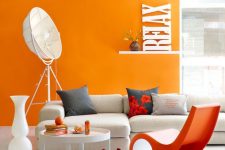 a bold contemporary living room with an orange accent wall, a white corner sofa, an orange chair, a round table, some quirky lamps