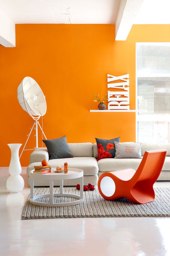 a bold contemporary living room with an orange accent wall, a white corner sofa, an orange chair, a round table, some quirky lamps