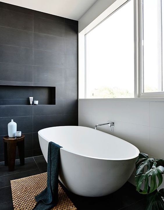 a bold modern bathroom clad with white and dark concrete tiles, with an oval tub, a mat, potted plants and a dark stained stool