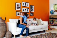 a bright living room with an orange accent wall, a white sofa, open storage units and a colorful rug plus a gallery wall with family pics
