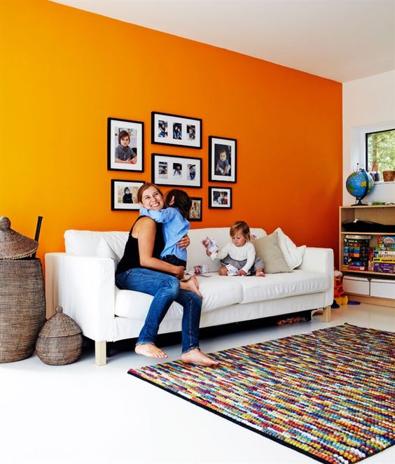 a bright living room with an orange accent wall, a white sofa, open storage units and a colorful rug plus a gallery wall with family pics