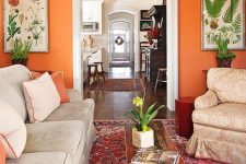 a bright living room with an orange accent wall, neutral vintage furniture, a printed rug, a low inlaid coffee table and botanical posters