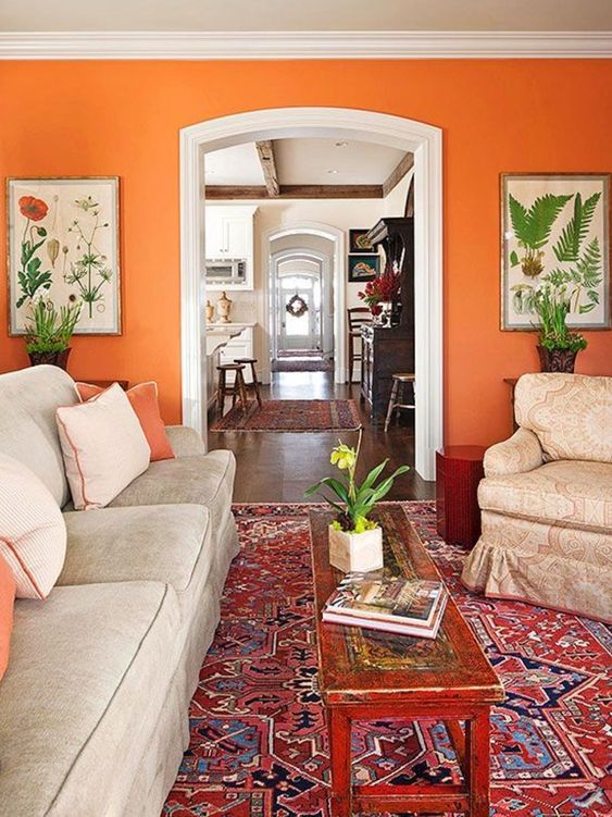 a bright living room with an orange accent wall, neutral vintage furniture, a printed rug, a low inlaid coffee table and botanical posters