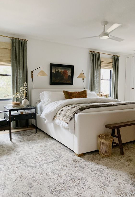 a chic and welcoming bedroom with a creamy upholstered bed, neutral bedding, chic nightstands, a wooden bench, baskets and green curtains