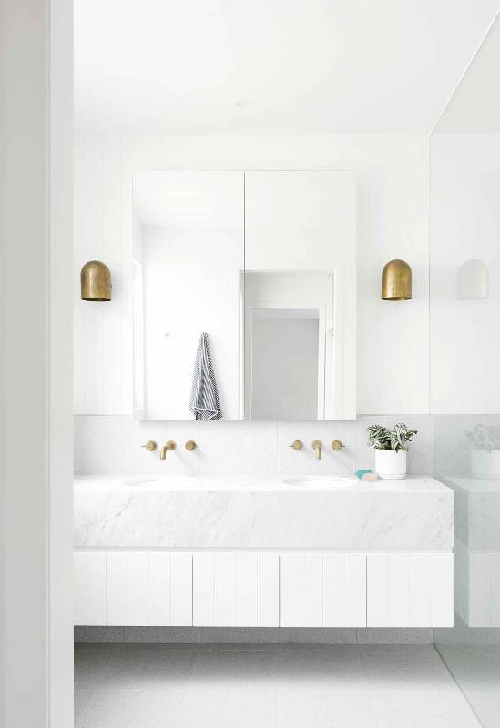 a clean white bathroom with a large mirror, a large floating vanity clad with marble and tiles and brass fixtures is cool