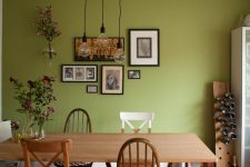 a contemporary dining room with a bold grene accent wall with a small gallery wall, pendant bulbs, a cool dining table and mismatching chairs