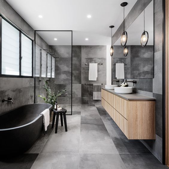 a contemporary grey bathroom clad with concrete tiles, a stone bathtub, a floating vanity, round sinks, black fixtures and framing