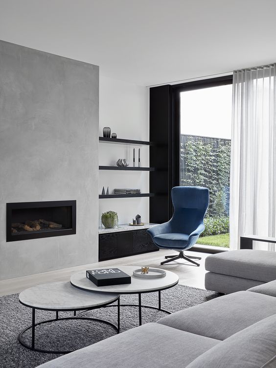 a contemporary living room with a concrete fireplace, built-in shelves, a blue chair, a grey sectional and a duo of tables