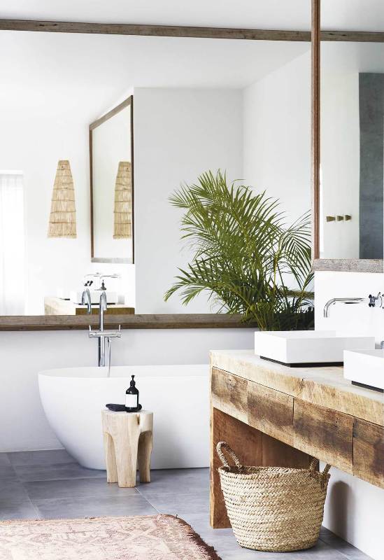 a contemporary tropical bathrom with a mirror that takes a whole wall, a wooden vanity, a tub, a basket and some potted plants